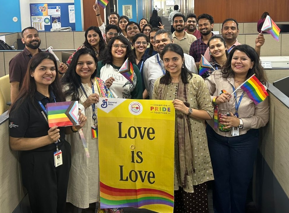 A group of cheerful people smiling and holding Rainbow flags, along with a banner that reads "Love is Love," featuring the GeneralMills logo in the top left corner. The scene depicts a celebration for the Mumbai Queer Azadi Month.